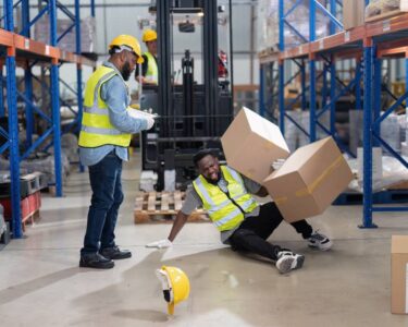 Two Black warehouse workers in safety vests. One worker fell over with two boxes falling on top of him.