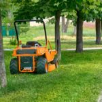 A commercial lawn mower is parked on the grass between several trees. The sidewalk has freshly cut grass scattered.