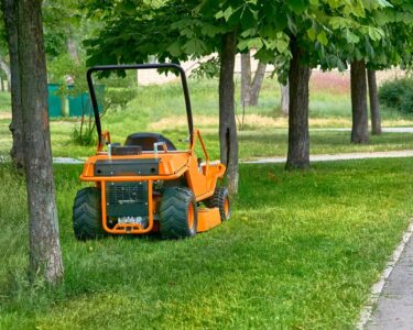 A commercial lawn mower is parked on the grass between several trees. The sidewalk has freshly cut grass scattered.