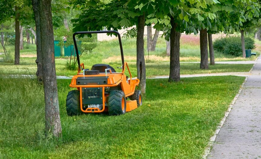 A commercial lawn mower is parked on the grass between several trees. The sidewalk has freshly cut grass scattered.