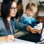 A woman at a desk working on a laptop and writing in a notebook while her concentrated son writes something next to her.