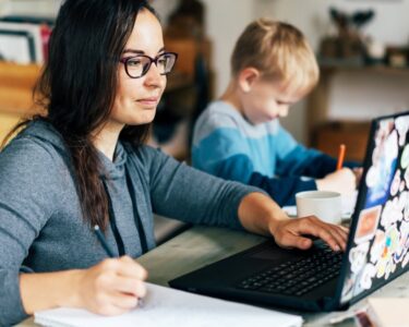 A woman at a desk working on a laptop and writing in a notebook while her concentrated son writes something next to her.