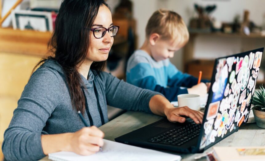 A woman at a desk working on a laptop and writing in a notebook while her concentrated son writes something next to her.