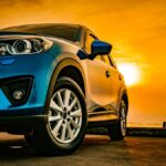 A close-up view shows a blue SUV parked on the beach, with the sun creating an orange sky in the background.