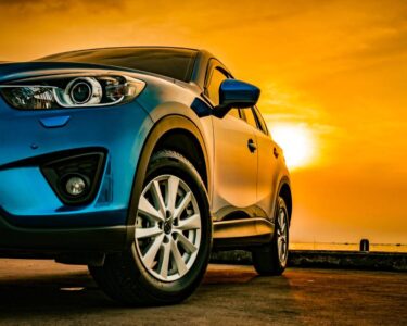 A close-up view shows a blue SUV parked on the beach, with the sun creating an orange sky in the background.