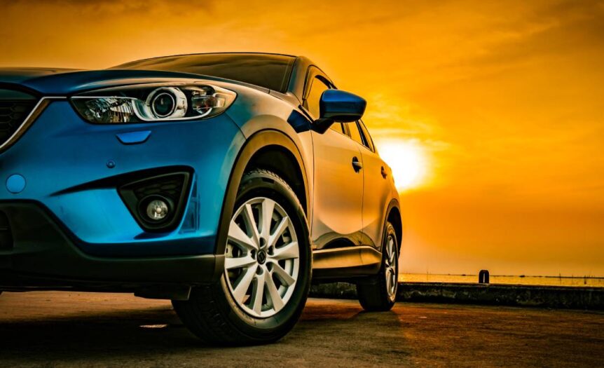 A close-up view shows a blue SUV parked on the beach, with the sun creating an orange sky in the background.