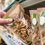 A person's hand grabbing the edge of a paper and plastic pouch of walnuts to pull it off a grocery store shelf.