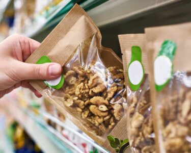 A person's hand grabbing the edge of a paper and plastic pouch of walnuts to pull it off a grocery store shelf.