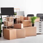 A bunch of stacked moving boxes surrounded by office chairs, shelves, plants, and monitors in an empty white room.