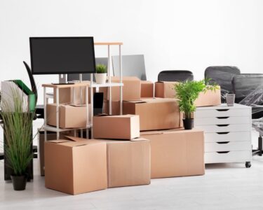 A bunch of stacked moving boxes surrounded by office chairs, shelves, plants, and monitors in an empty white room.