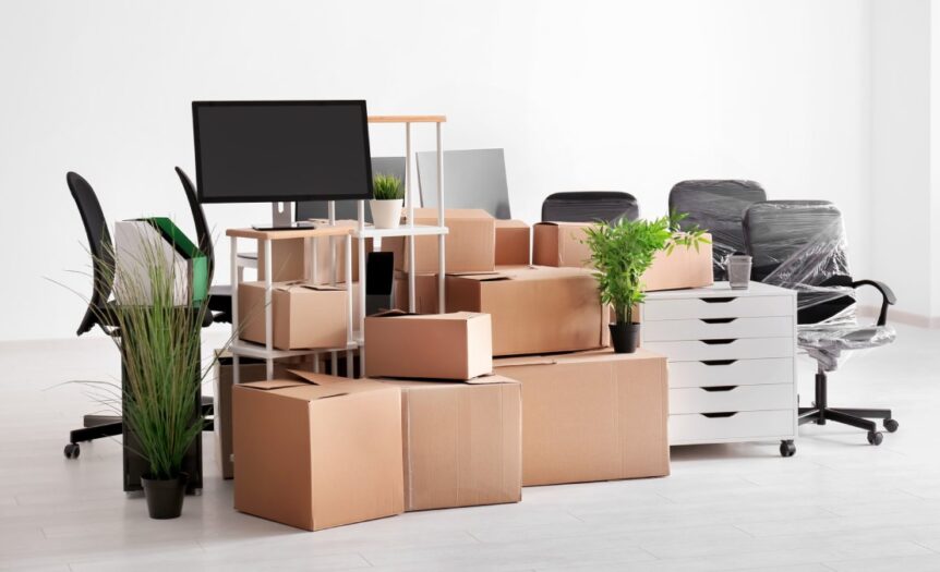 A bunch of stacked moving boxes surrounded by office chairs, shelves, plants, and monitors in an empty white room.