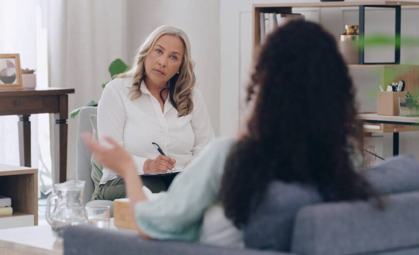 A woman sits in a gray chair, writing on a clipboard and intently looking at a woman sitting on a blue couch.