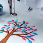 A close-up view shows an embroidery machine embroidering a brown tree with blue and pink leaves onto a white fabric.