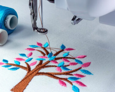 A close-up view shows an embroidery machine embroidering a brown tree with blue and pink leaves onto a white fabric.