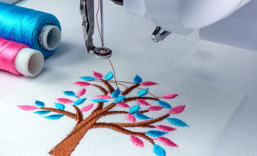 A close-up view shows an embroidery machine embroidering a brown tree with blue and pink leaves onto a white fabric.