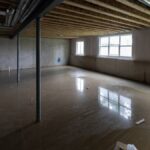 A large unfinished basement with several windows and an exposed ceiling has a layer of water on the concrete floor.