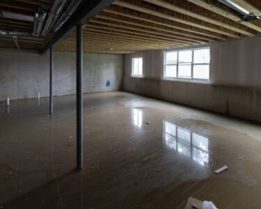 A large unfinished basement with several windows and an exposed ceiling has a layer of water on the concrete floor.