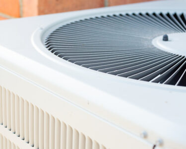 A close-up of the top half of a clean, outdoor HVAC unit. An orange brick wall is in the background of the AC condenser.