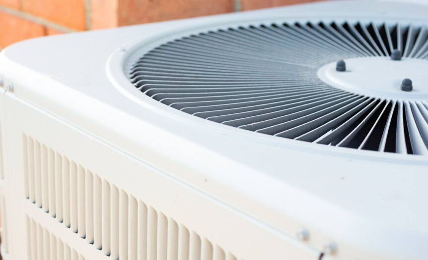 A close-up of the top half of a clean, outdoor HVAC unit. An orange brick wall is in the background of the AC condenser.