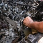 Two pairs of hands, one gloved and holding a screwdriver, are working on an exposed diesel truck engine.