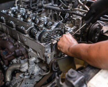 Two pairs of hands, one gloved and holding a screwdriver, are working on an exposed diesel truck engine.