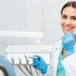 A dental professional holds her mask down to show off a smile while sitting in front of a patient and holding a tool.