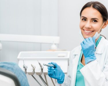 A dental professional holds her mask down to show off a smile while sitting in front of a patient and holding a tool.