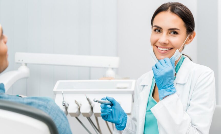 A dental professional holds her mask down to show off a smile while sitting in front of a patient and holding a tool.