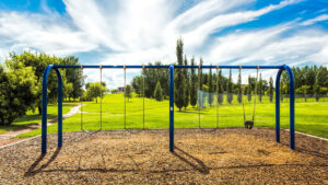 A blue swing set in an empty park built on a patch of mulch. The swing set is surrounded by lush, green grass.