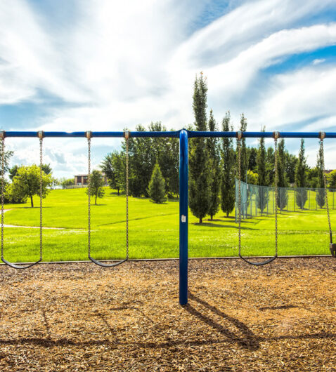 A blue swing set in an empty park built on a patch of mulch. The swing set is surrounded by lush, green grass.