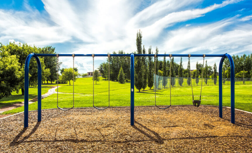 A blue swing set in an empty park built on a patch of mulch. The swing set is surrounded by lush, green grass.