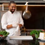 A chef in a white uniform standing in a kitchen surrounded by ingredients as he slips on his gloves for safety.