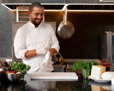 A chef in a white uniform standing in a kitchen surrounded by ingredients as he slips on his gloves for safety.