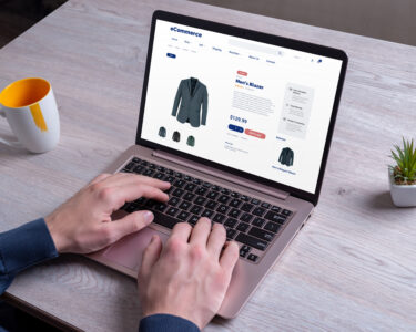 A person sitting at the table on their laptop browsing an online e-commerce site. They have a shirt pulled up on the screen.