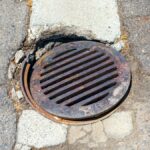 A broken manhole cover in the middle of the road. The cover is rusting and the hole in the ground is eroding.