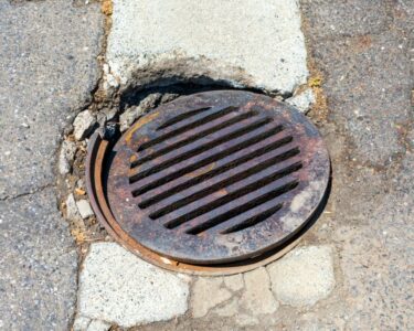 A broken manhole cover in the middle of the road. The cover is rusting and the hole in the ground is eroding.