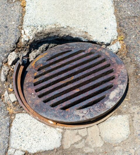 A broken manhole cover in the middle of the road. The cover is rusting and the hole in the ground is eroding.