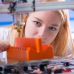 A young woman stands behind a 3D printer, adjusting an orange-colored plastic part manufactured by the machine.