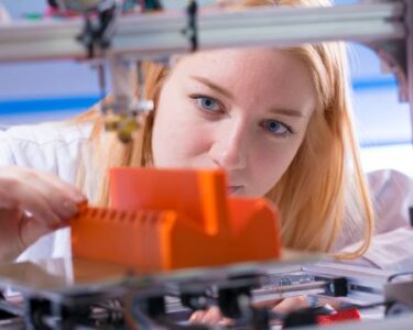 A young woman stands behind a 3D printer, adjusting an orange-colored plastic part manufactured by the machine.