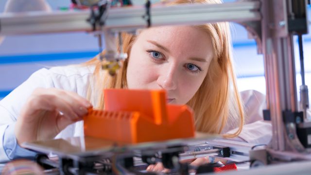 A young woman stands behind a 3D printer, adjusting an orange-colored plastic part manufactured by the machine.