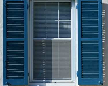 Closeup of a window on a white stone house with bright blue exterior shutters that are pushed open on both sides.