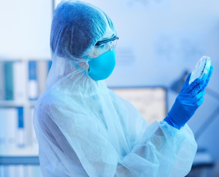 A woman wearing a white gown, safety glasses, a mask, and gloves is examining the contents of a petri dish.