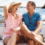 A laughing older couple sits close together on the cushioned back seat of a boat with pillows. A wake trails behind them.