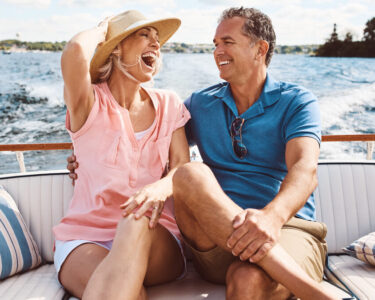 A laughing older couple sits close together on the cushioned back seat of a boat with pillows. A wake trails behind them.