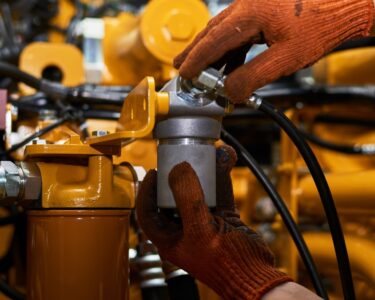 A pair of hands wearing dark orange work gloves holding hydraulic system components. There is orange machinery in the background.