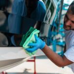 A young man wearing a white T-shirt and blue gloves uses a green washcloth to clean the side of a boat.