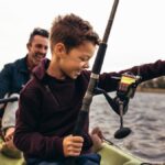 A man and small child fish inside a green kayak. The child is smiling and pulling up his fishing line.