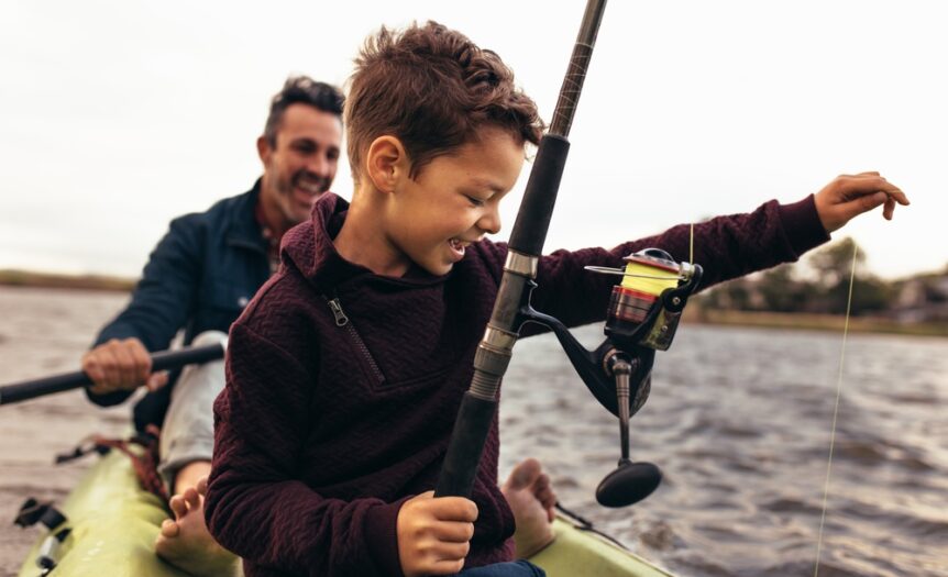 A man and small child fish inside a green kayak. The child is smiling and pulling up his fishing line.