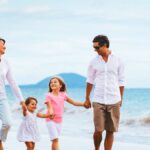 A mother, father, and two young daughters walk smiling, hand in hand, along the shore of a tropical beach.