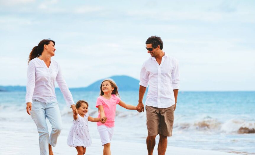 A mother, father, and two young daughters walk smiling, hand in hand, along the shore of a tropical beach.